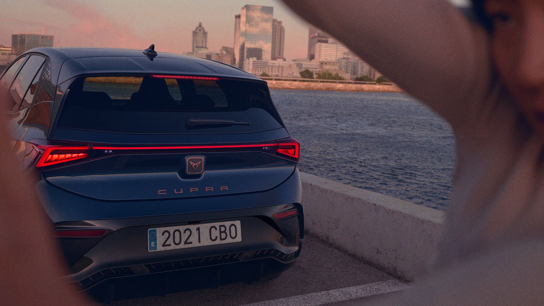 Moder CUPRA Born je parkiran ob reki z mestno pokrajino v ozadju. Oseba z zamegljenim obrazom je delno vidna na desni strani slike.