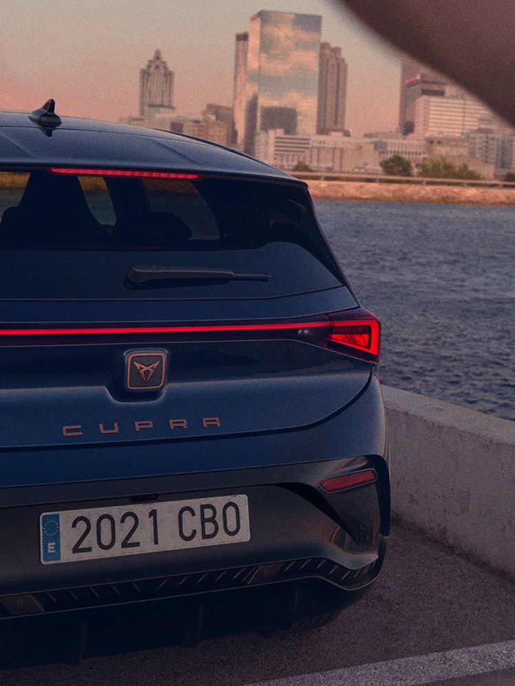 Moder CUPRA Born je parkiran ob reki z mestno pokrajino v ozadju. Oseba z zamegljenim obrazom je delno vidna na desni strani slike.