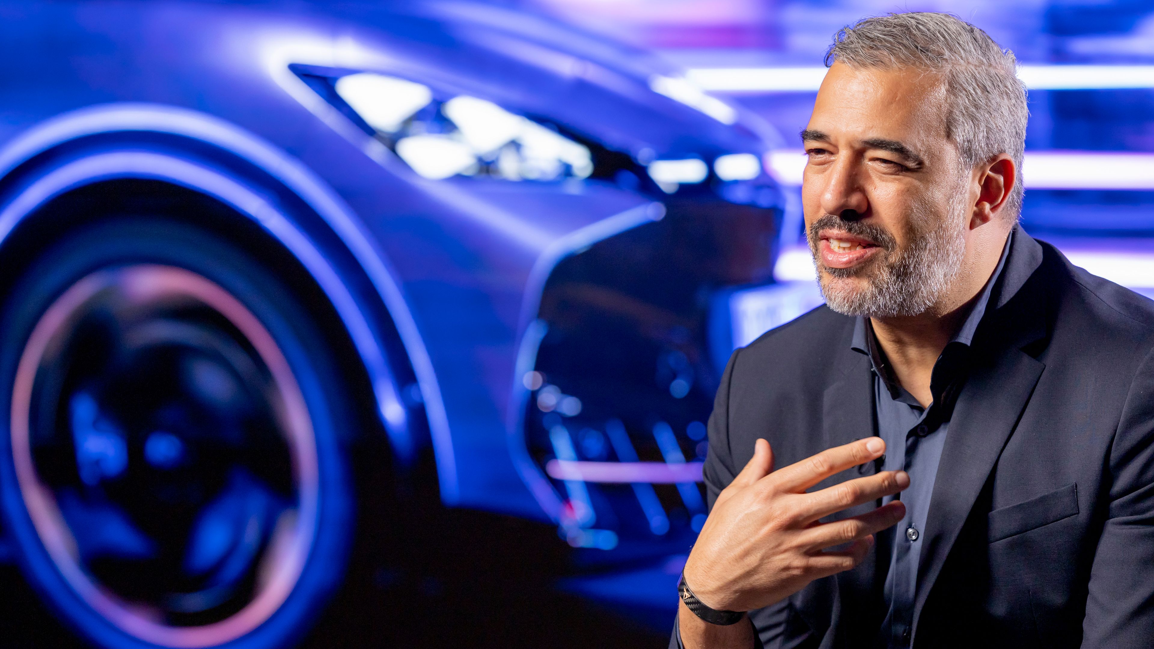 Oblikovalec Jorge Díez sedi pred zamegljeno svetlečo podobo avtomobila, oblečen je v poslovno obleko in se zdi, da gestikulira med govorom.