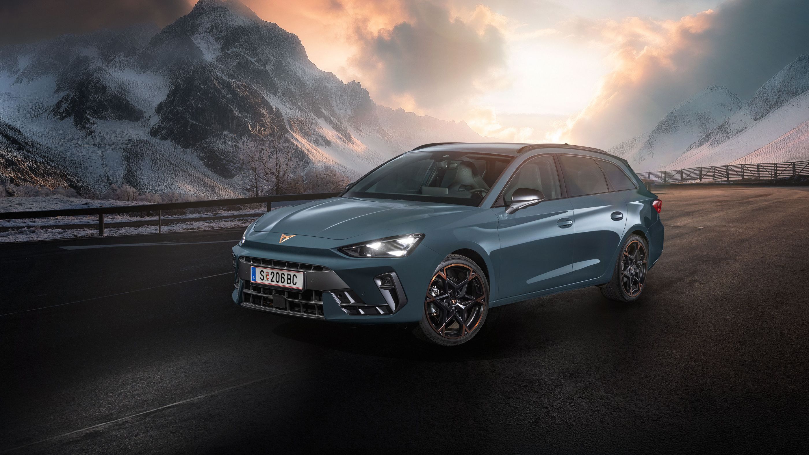 Ein CUPRA Leon Kombi parkt vor einer malerischen Berglandschaft bei Sonnenuntergang. Im Hintergrund sind verschneite Gipfel und dramatische Wolken zu sehen. Der Asphalt ist dunkel und feucht.