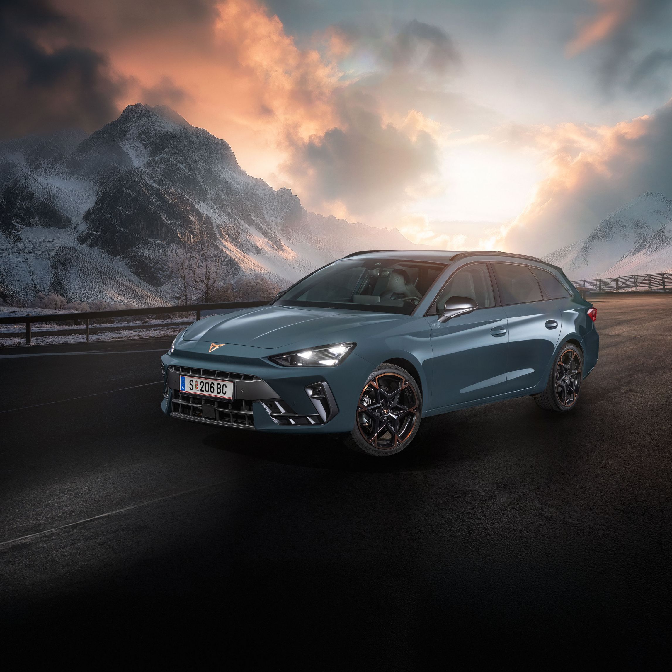 Ein CUPRA Leon Kombi parkt vor einer malerischen Berglandschaft bei Sonnenuntergang. Im Hintergrund sind verschneite Gipfel und dramatische Wolken zu sehen. Der Asphalt ist dunkel und feucht.