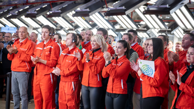 Skupina ljudi, oblečenih v rdeče uniforme, stoji in ploska v zaprtem prostoru, ki ima veliko luči na stropu. 