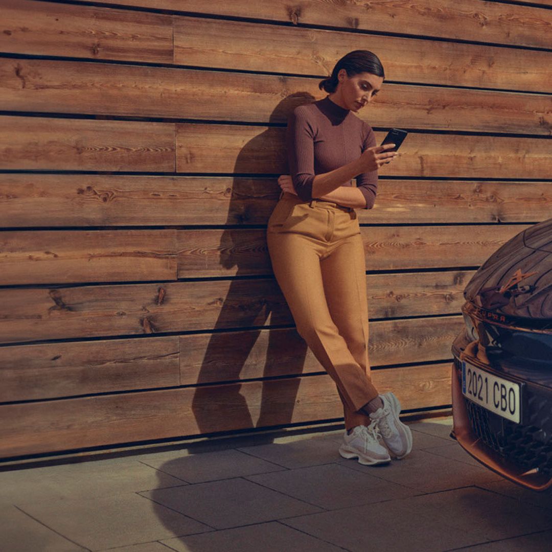 Oseba stoji ob leseni steni in uporablja aplikacijo CUPRA za model Born. V bližini je parkiran moder CUPRA Born.