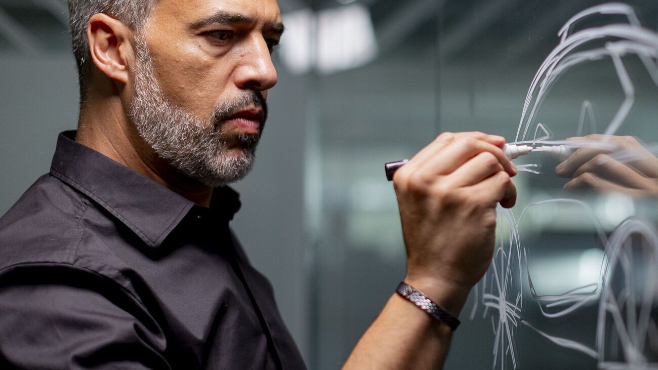 Moški riše s suho piši markerjem po stekleni tabli, na kateri so skice različnih oblik. 