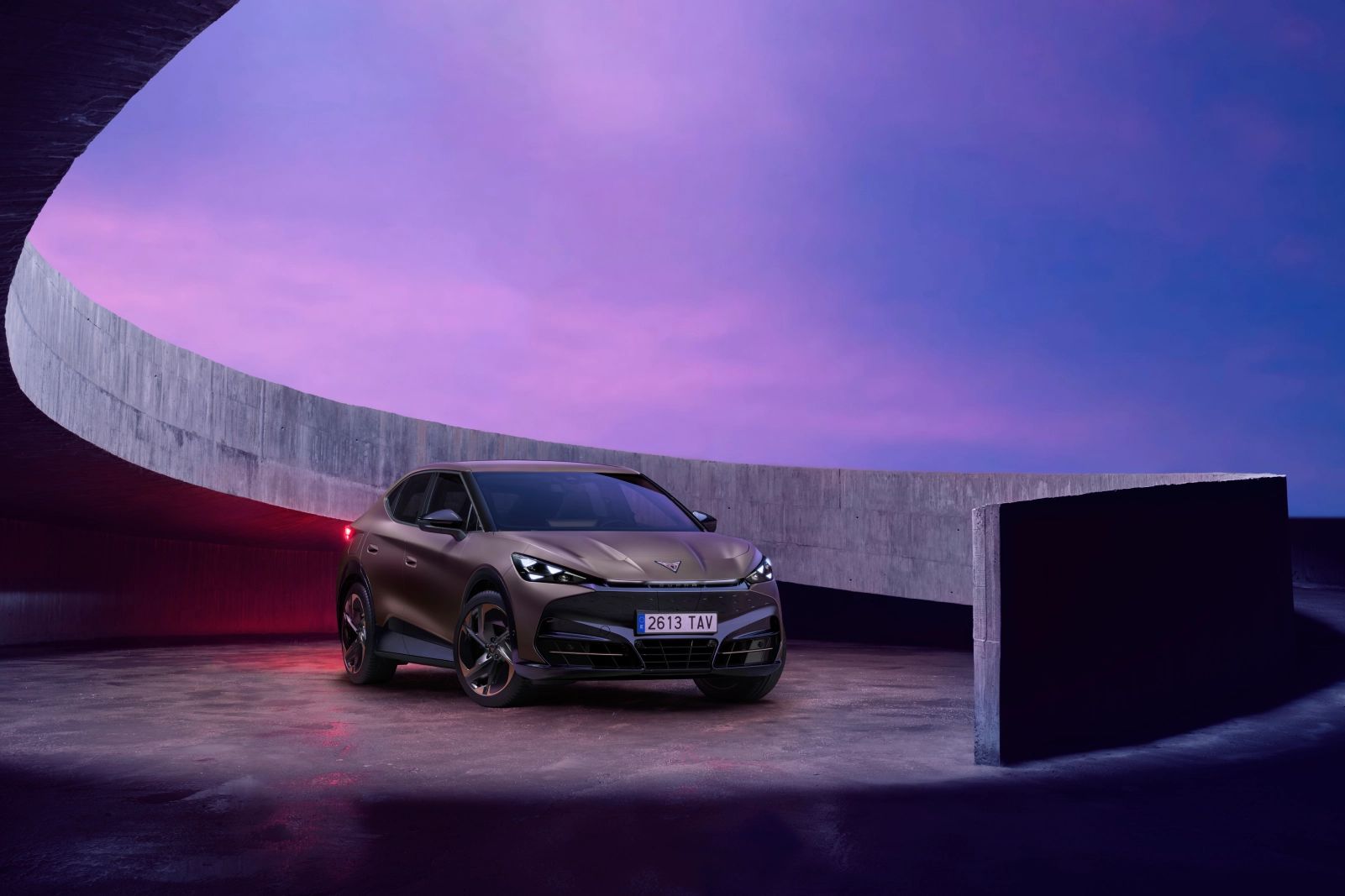 CUPRA Tavascan stoji na betonski rampi z elegantnim, ukrivljenim zidom. Ozadje je osvetljeno z dramatičnim vijoličnim in rožnatim nebom.