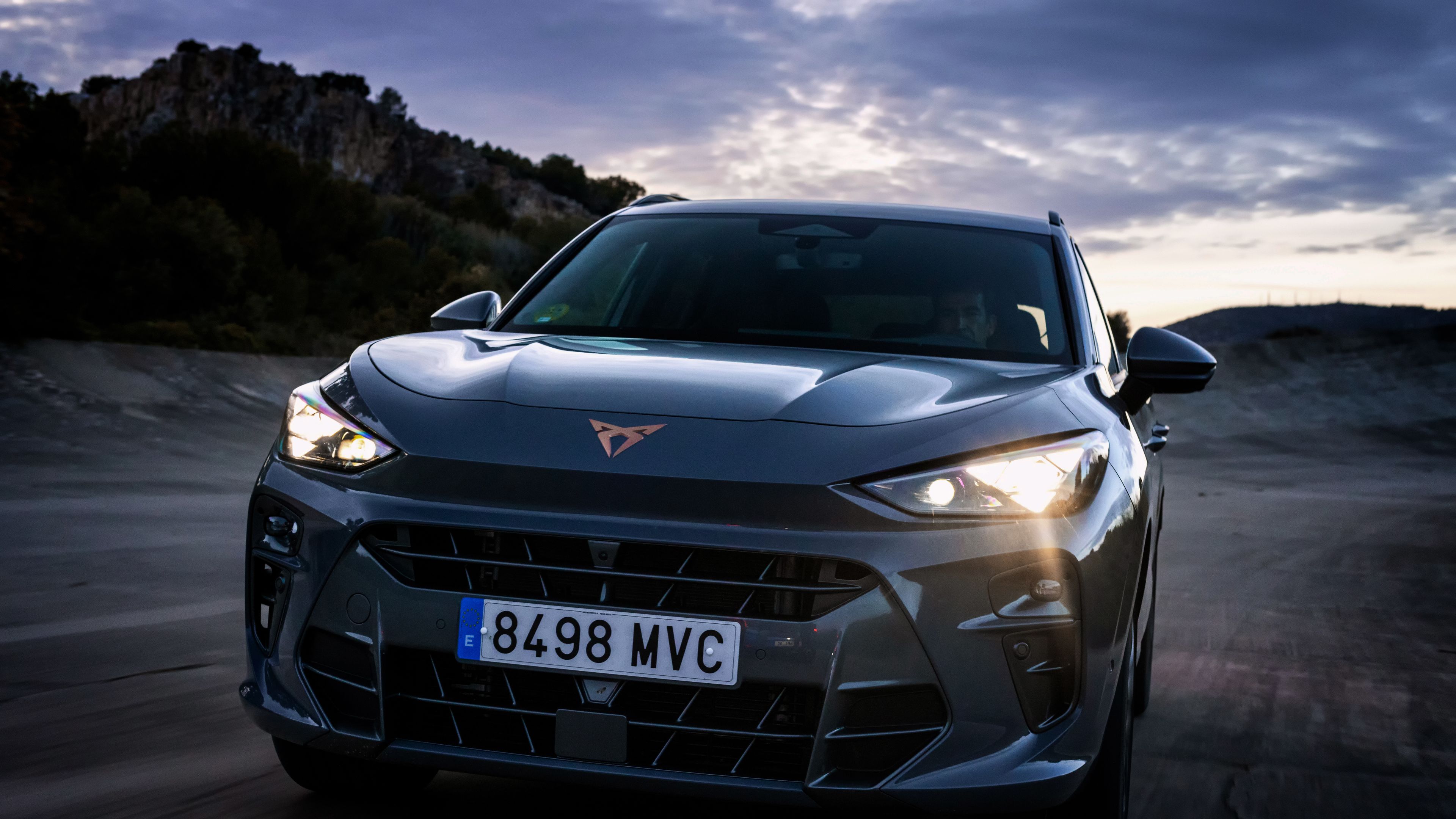 CUPRA Terramar vozi po asfaltni cesti ob mraku, s prižganimi žarometi, v ozadju pa so hribi in oblačno nebo.