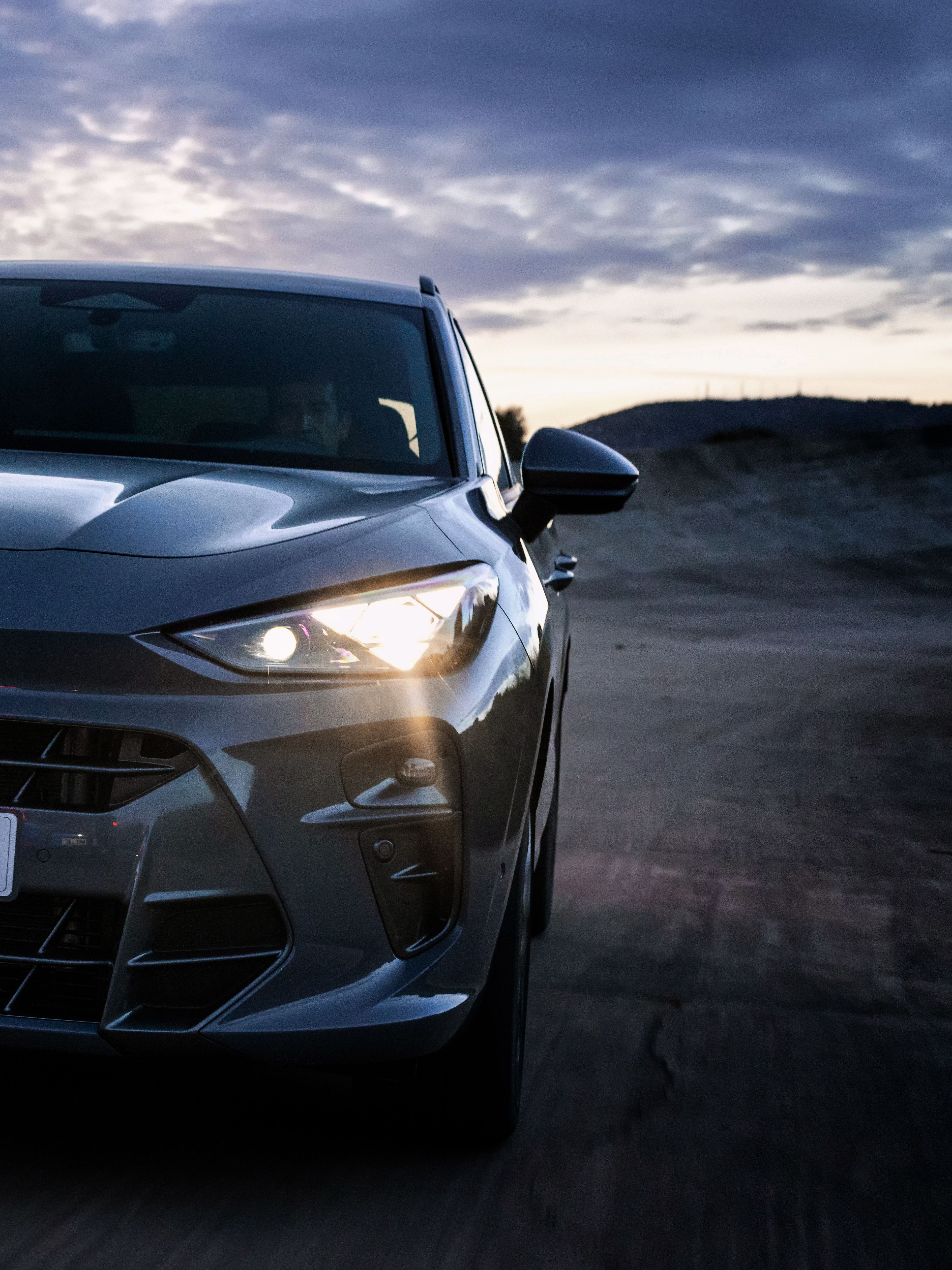 CUPRA Terramar vozi po asfaltni cesti ob mraku, s prižganimi žarometi, v ozadju pa so hribi in oblačno nebo.