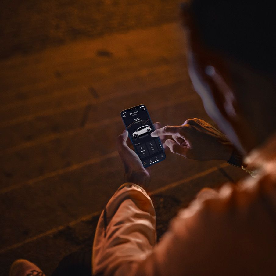 Oseba sedi na stopnicah in uporablja aplikacijo 'My CUPRA app' na pametnem telefonu. Aplikacija prikazuje podobe avtomobila. Osvetlitev je temna, s toplim sijajem.
