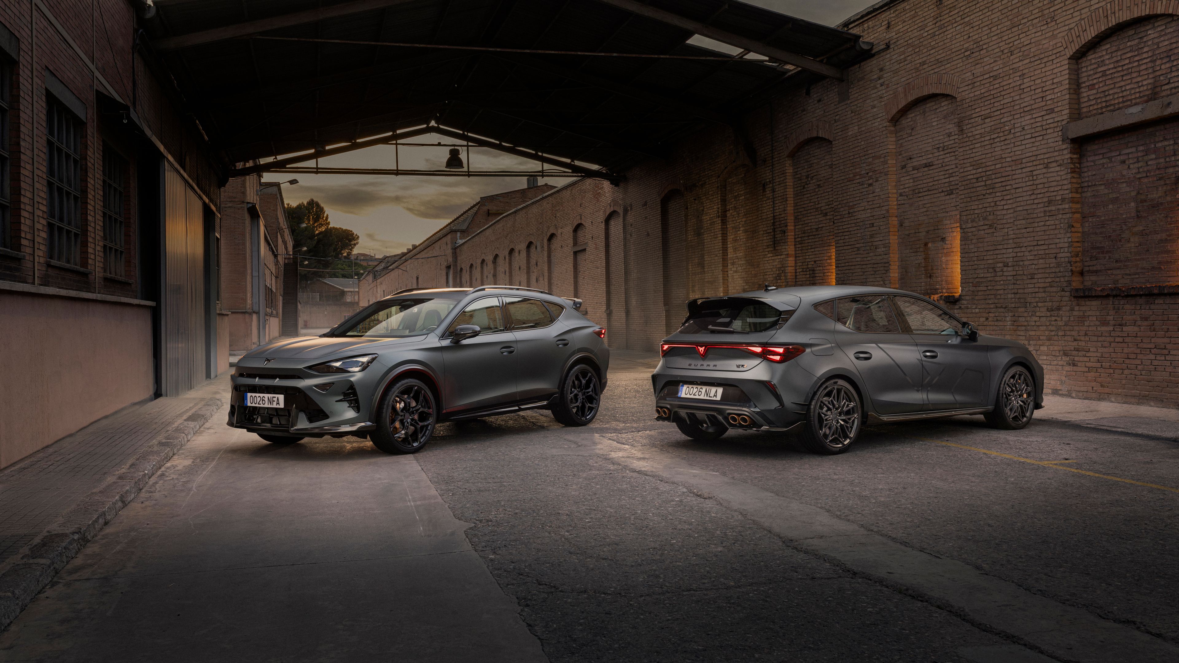 Dve športni sivi vozili CUPRA Formentor in CUPRA Leon sta parkirani na ozki industrijski ulici med starimi opečnatimi zgradbami ob mraku.