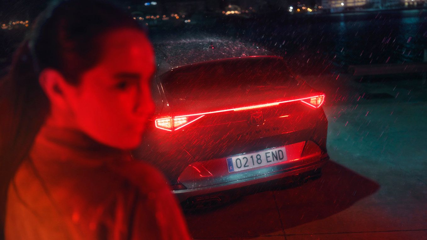 Ženska stoji pred avtomobilom CUPRA Formentor, ki ima prižgane zadnje luči. Scena je ponoči in rahlo deževna.