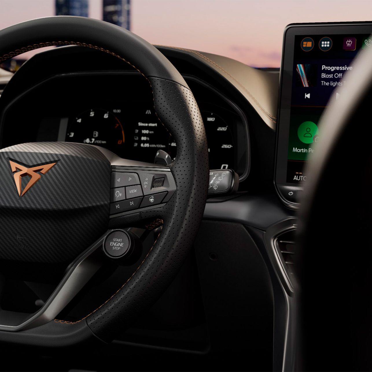 Volan avtomobila Cupra Leon z logotipom znamke Cupra, obdano z perforiranim usnjem in oranžnimi šivi. Na volanskem obroču so vidni gumbi za upravljanje. V ozadju je vidna armaturna plošča in zaslon na dotik.