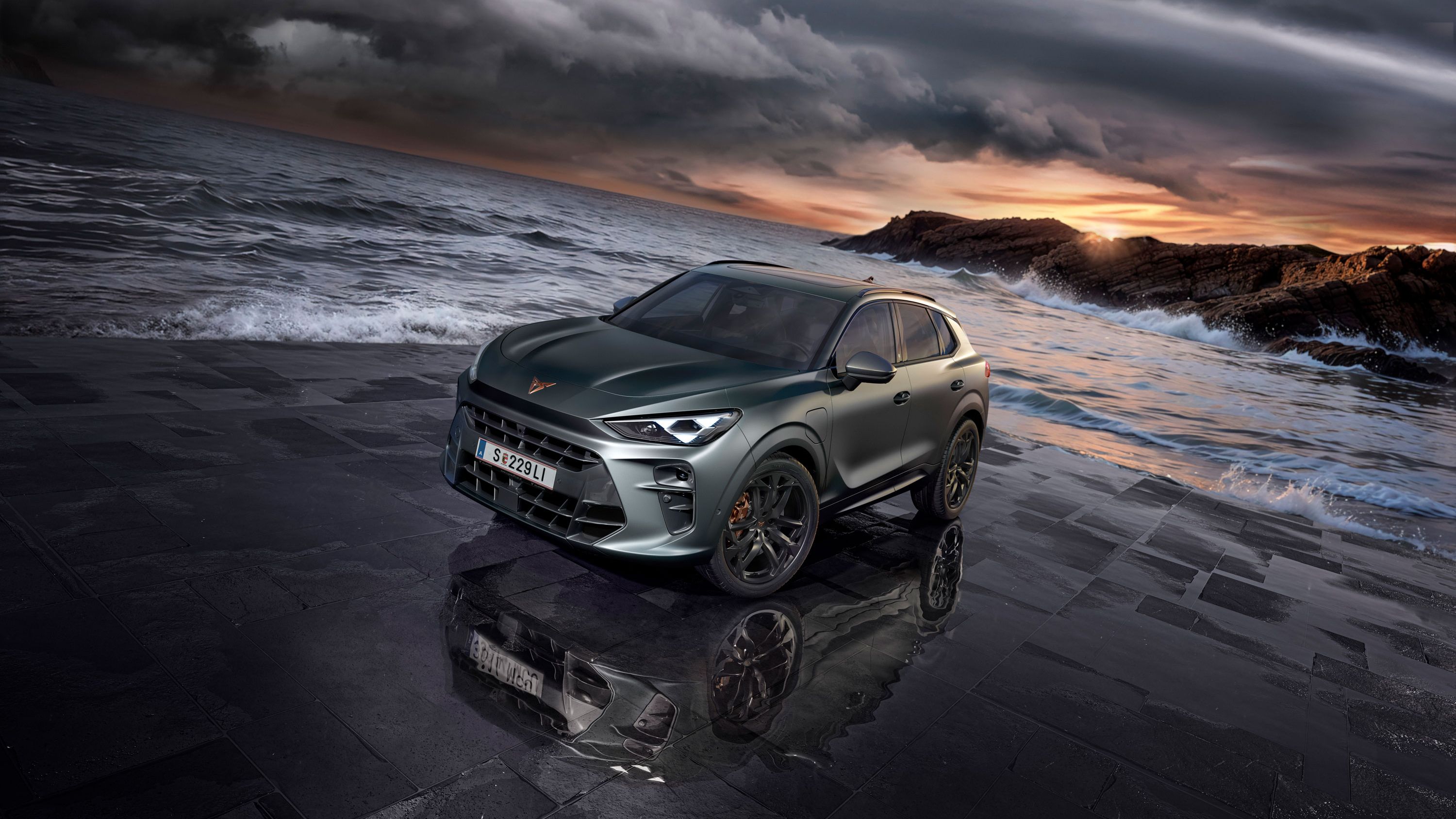 Sodobna temno sivo vozilo CUPRA Terramar, s črnimi platišči, na mokrem tlakovcu ob obali z oblačnim nebom v ozadju. Morje, valovi in skalnata obala se raztezajo v ozadju.