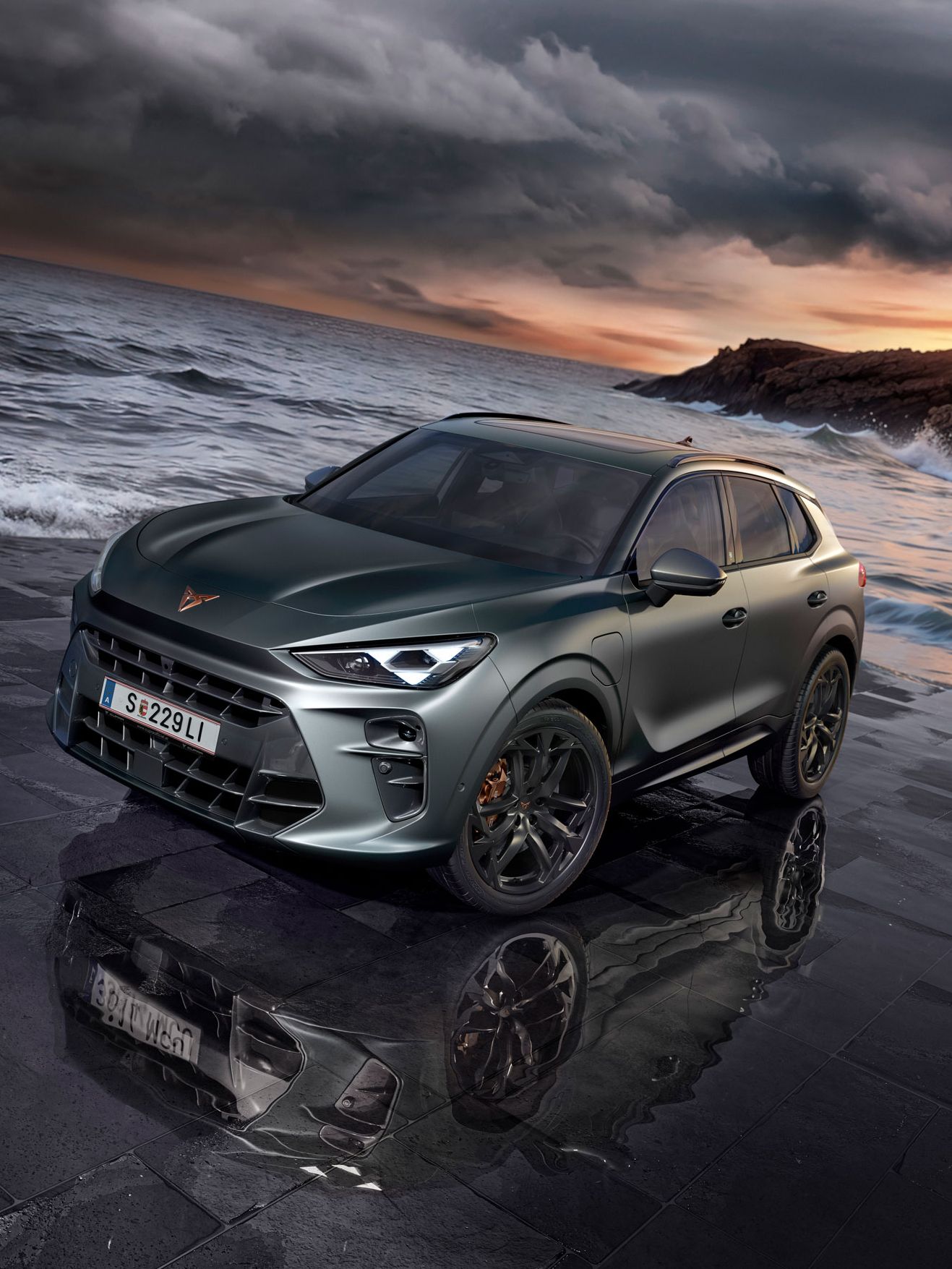 Sodobna temno sivo vozilo CUPRA Terramar, s črnimi platišči, na mokrem tlakovcu ob obali z oblačnim nebom v ozadju. Morje, valovi in skalnata obala se raztezajo v ozadju.