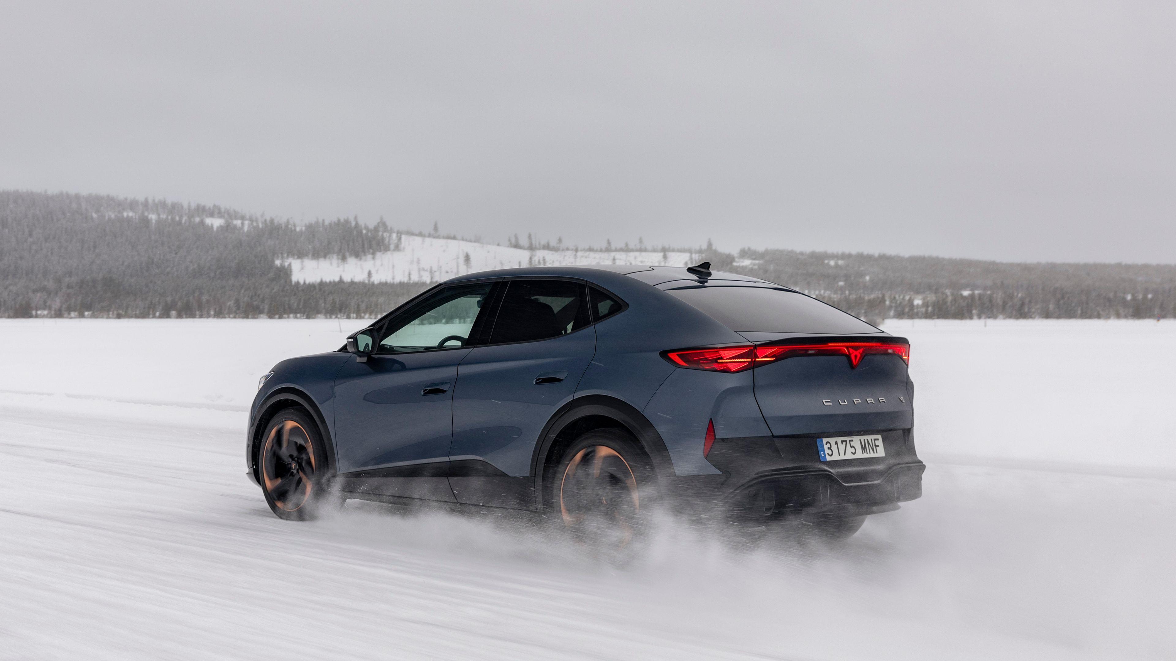 Temno modri CUPRA Tavascan vozi po zasneženi površini v zimskem okolju. Zadnji del vozila je dobro viden in ima sodoben SUV dizajn.