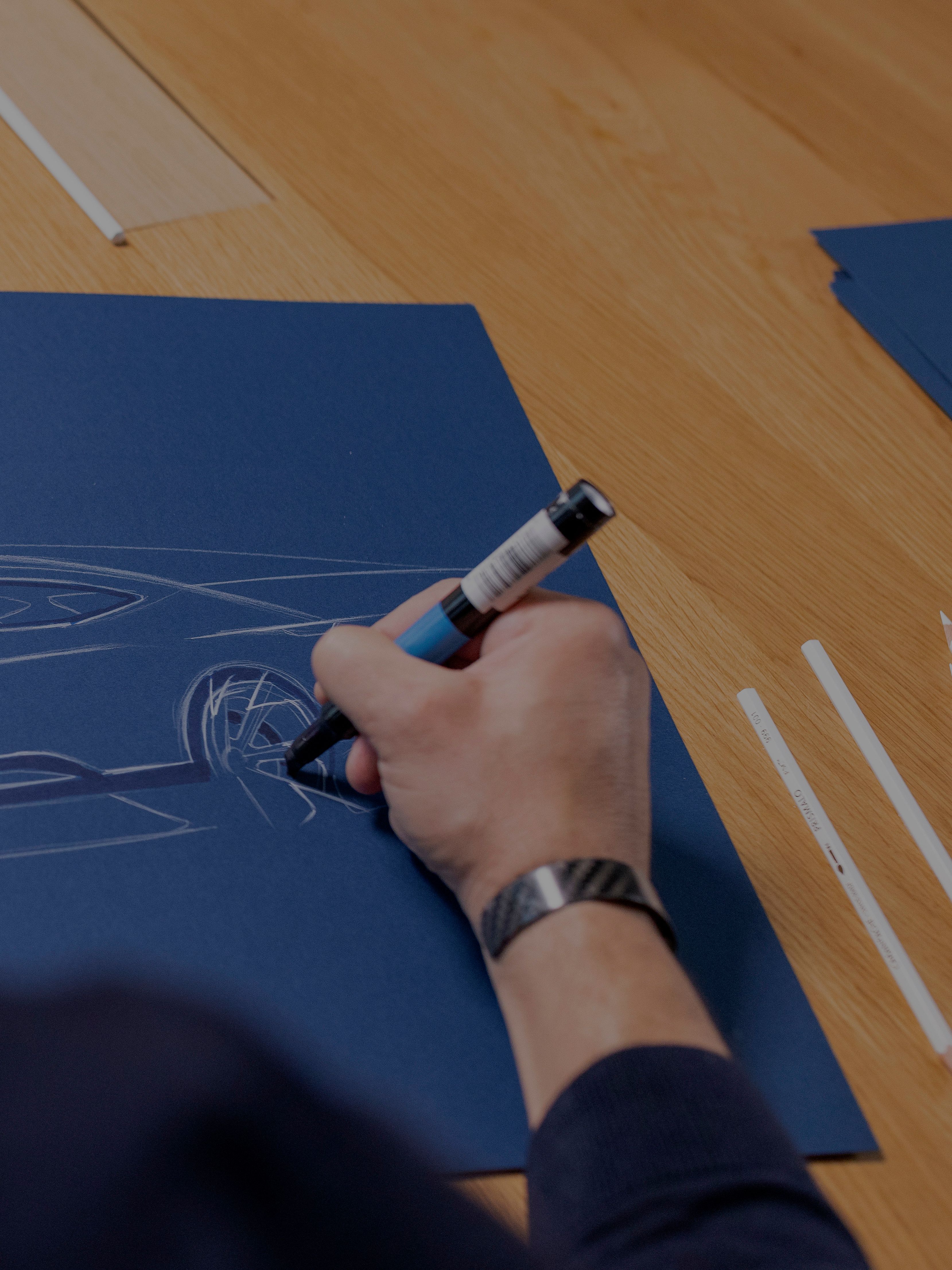 Oblikovalec riše skico avtomobila na modrem papirju. Risba prikazuje profil športnega avtomobila znamke CUPRA.