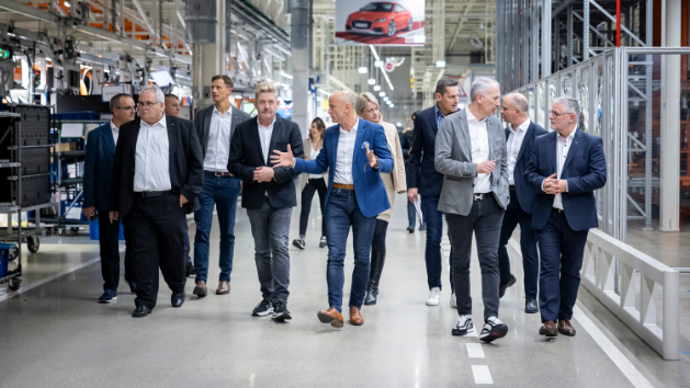 Skupina moških v poslovnih oblačilih se sprehaja po sodobni tovarniških prostorih, ki je polna industrijske opreme. V ozadju je oglas z avtomobilom.