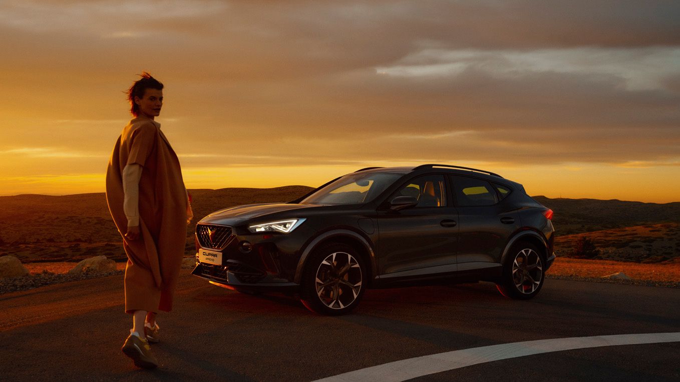 Ženska, hodi poleg parkiranega temno sivega vozila CUPRA Formentor ob sončnem zahodu. Vozilo je na cesti, v ozadju pa je vidna pokrajina.