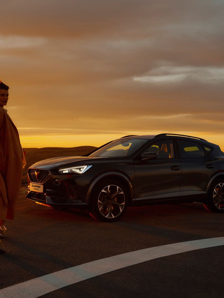 Ženska, hodi poleg parkiranega temno sivega vozila CUPRA Formentor ob sončnem zahodu. Vozilo je na cesti, v ozadju pa je vidna pokrajina.