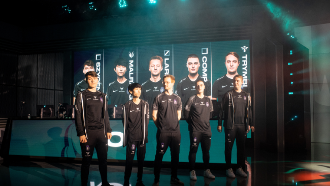 Pet igralcev v črnih dresih stoji na odru pred velikim zaslonom, ki prikazuje njihova imena in slike. Osvetlitev je dramatična in svetlobni efekti dodajo vzdušje posebnega dogodka. V ozadju so vidni računalniki in oprema.