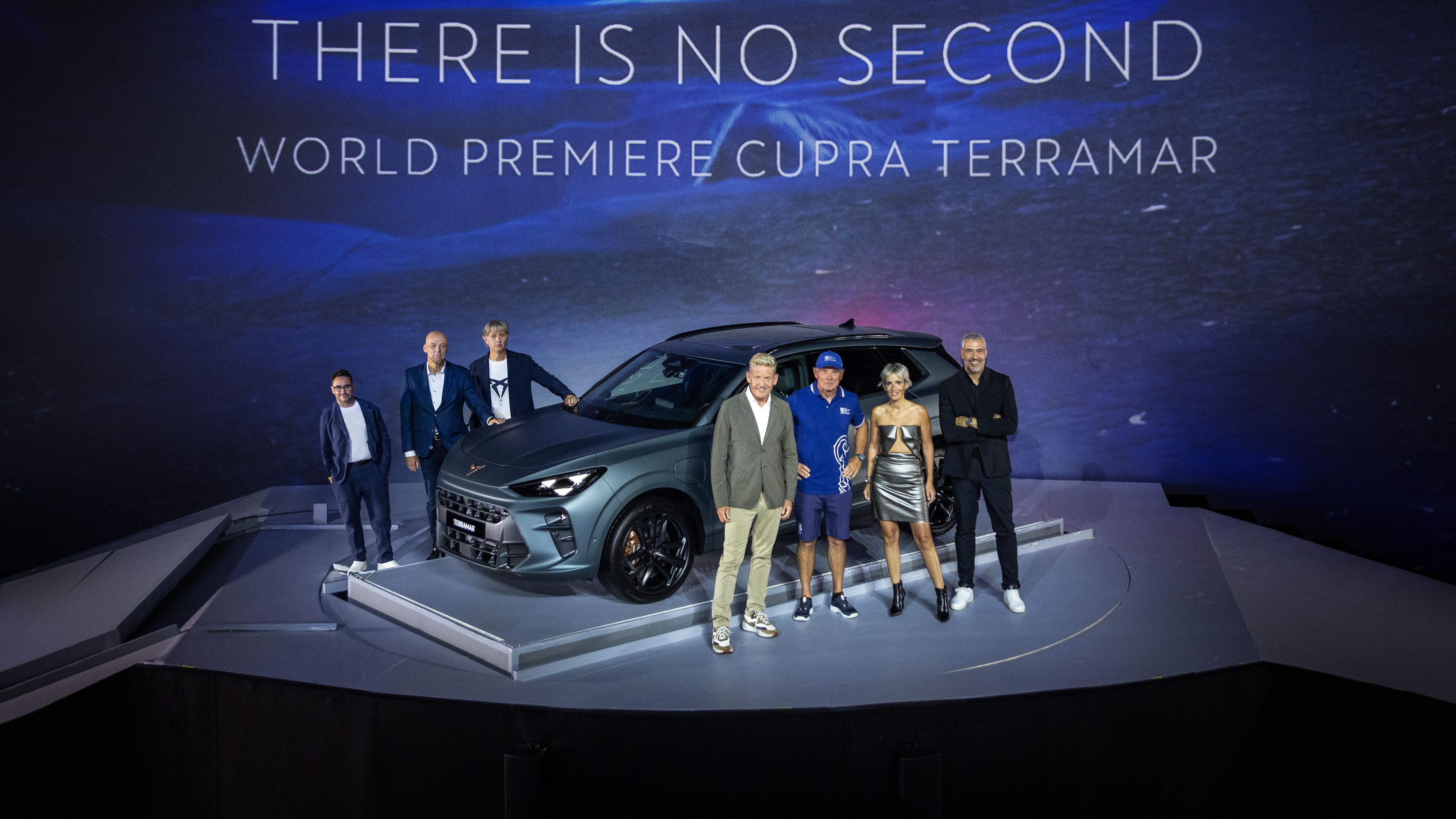 Na sliki je skupina ljudi, ki stojijo pred novim avtomobilom Cupra Terramar na svetovni premieri. Ozadje prikazuje modro osvetljeno grafiko z napisom "THERE IS NO SECOND world premiere CUPRA Terramar". 
