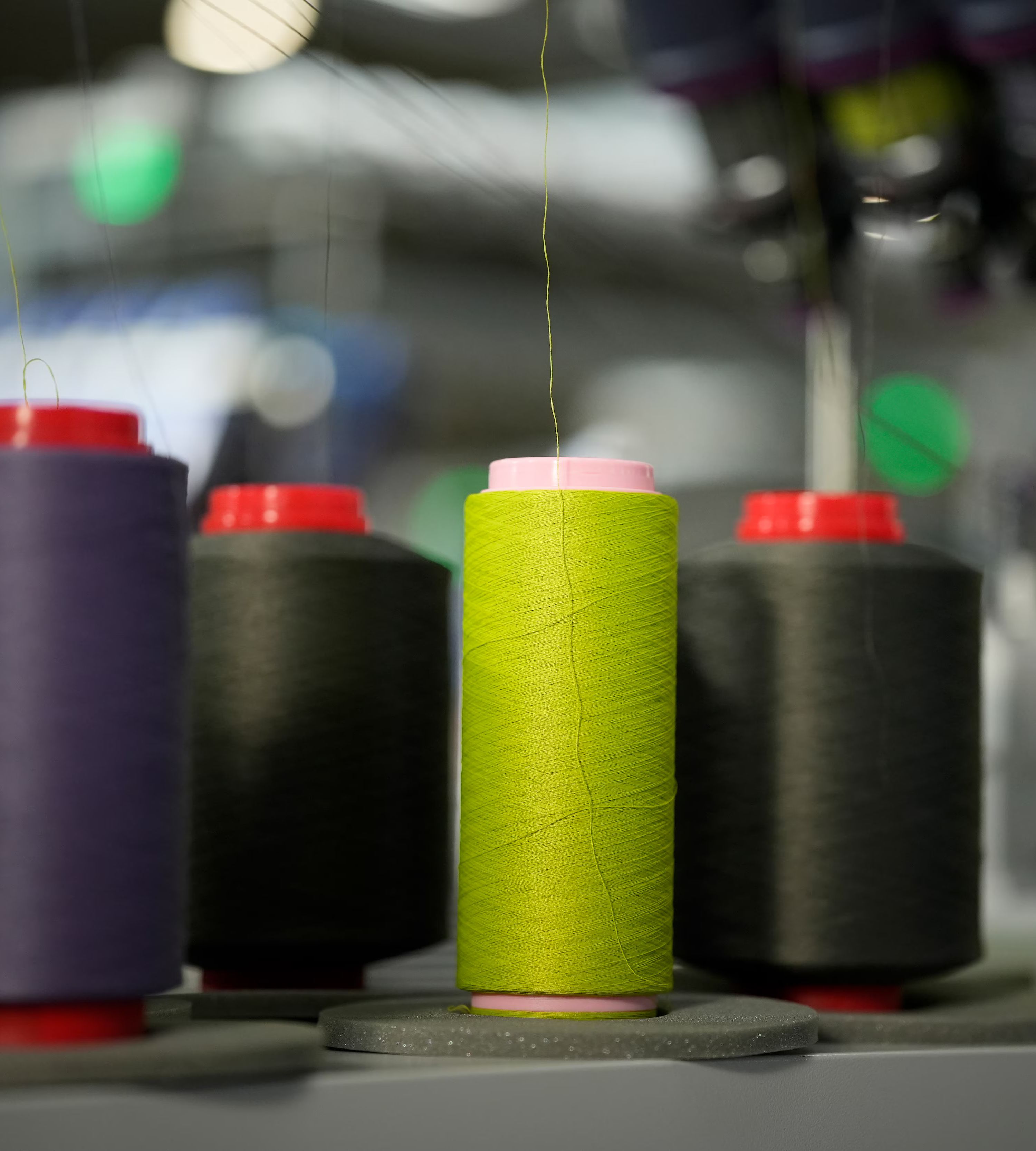 Tulci niti različnih barv stojijo na industrijskem stroju za izdelavo tekstila. V ospredju je poudarjena svetlo zelena nit.