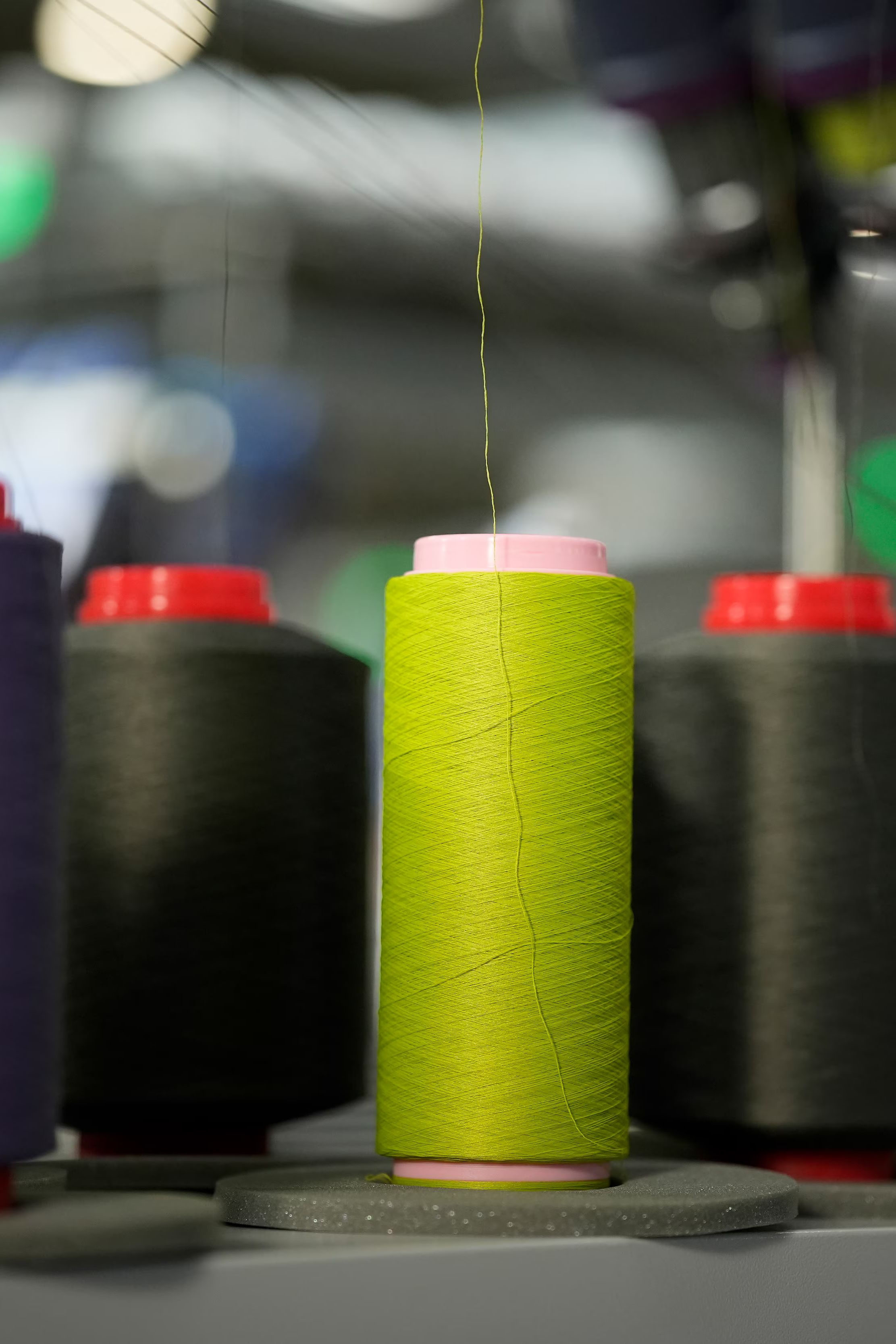 Tulci niti različnih barv stojijo na industrijskem stroju za izdelavo tekstila. V ospredju je poudarjena svetlo zelena nit.
