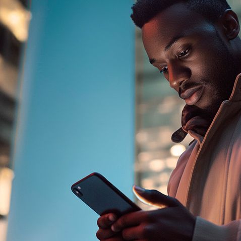 Oseba stoji na ulici v modernem mestnem okolju in gleda aplikacijo 'My CUPRA app' na pametnem telefonu. Okoli njega so visoke zgradbe z osvetljenimi okni, kar daje urbano nočno vzdušje. 