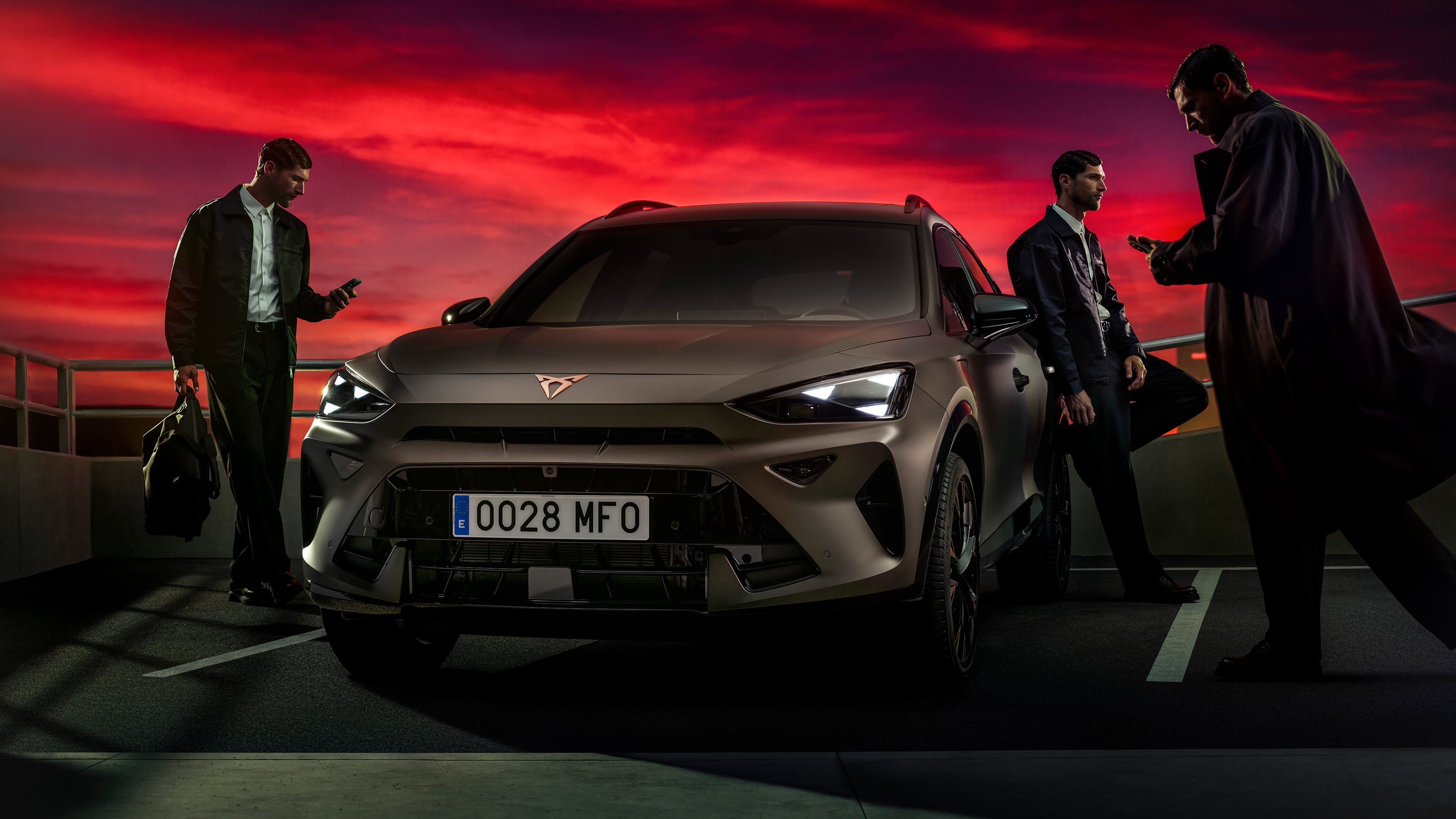 Bronasti CUPRA Formentor PA je parkiran na strešnem parkirišču ob dramatičnem rdečem sončnem zahodu, ob njem pa stojijo trije moški, ki gledajo v svoje telefone.