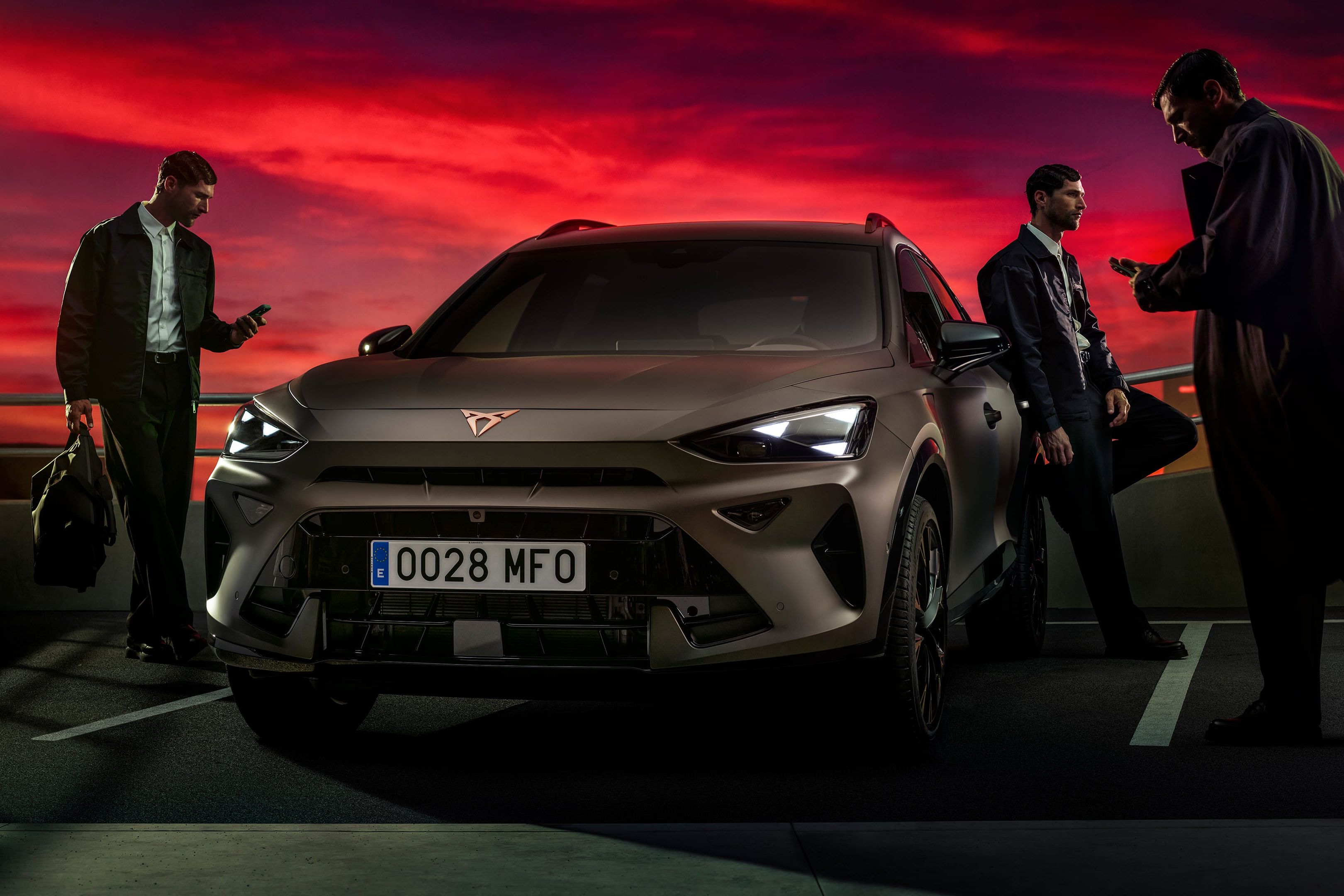 Bronasti CUPRA Formentor PA je parkiran na strešnem parkirišču ob dramatičnem rdečem sončnem zahodu, ob njem pa stojijo trije moški, ki gledajo v svoje telefone.