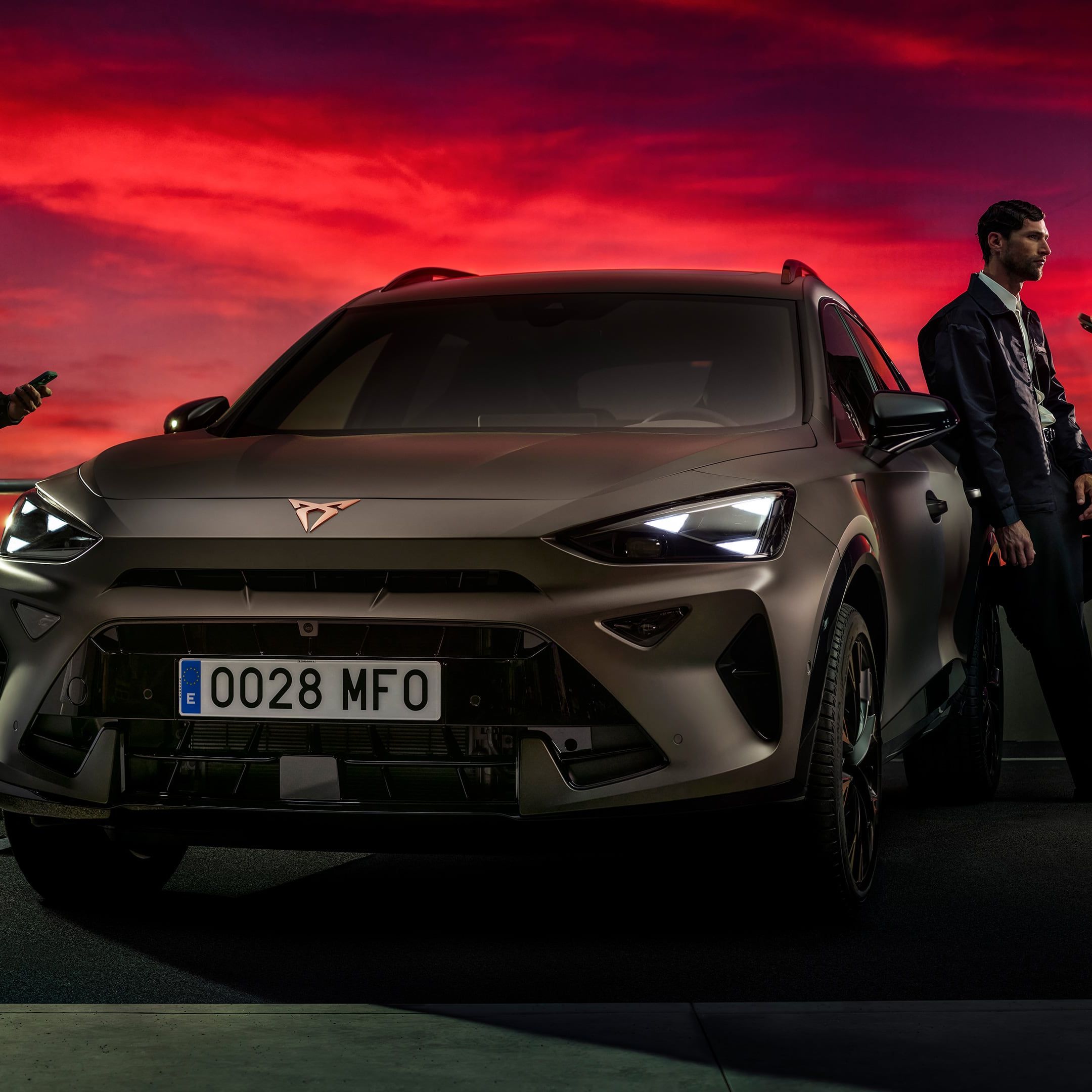 Bronasti CUPRA Formentor PA je parkiran na strešnem parkirišču ob dramatičnem rdečem sončnem zahodu, ob njem pa stojijo trije moški, ki gledajo v svoje telefone.