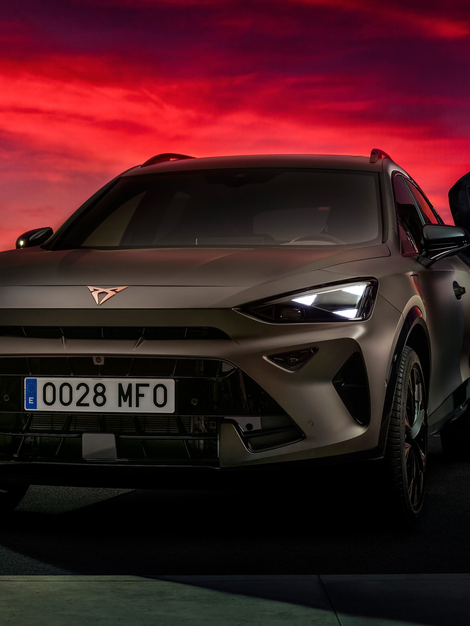 Bronasti CUPRA Formentor PA je parkiran na strešnem parkirišču ob dramatičnem rdečem sončnem zahodu, ob njem pa stojijo trije moški, ki gledajo v svoje telefone.