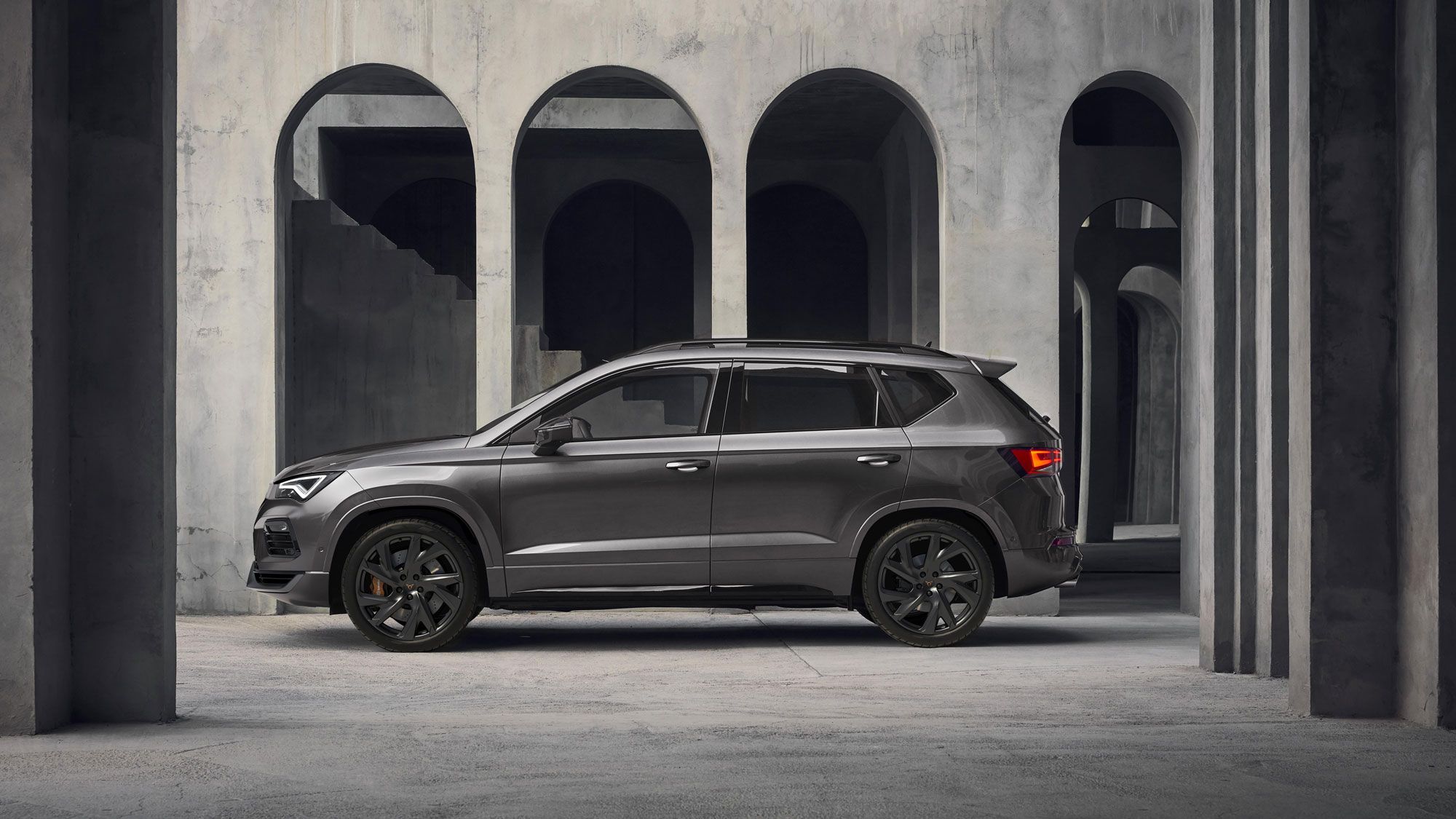 Sodoben CUPRA Ateca PA Lifestyle temne sive barve je parkiran v betonskem prostoru z masivnimi stebri in oboki. Avto je postavljen v stranski profil, kar poudarja njegovo elegantno obliko. Ozadje je minimalistično in industrijsko.