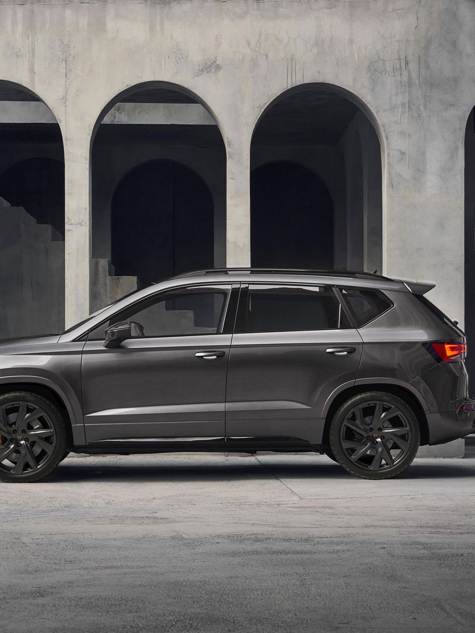 Sodoben CUPRA Ateca PA Lifestyle temne sive barve je parkiran v betonskem prostoru z masivnimi stebri in oboki. Avto je postavljen v stranski profil, kar poudarja njegovo elegantno obliko. Ozadje je minimalistično in industrijsko.