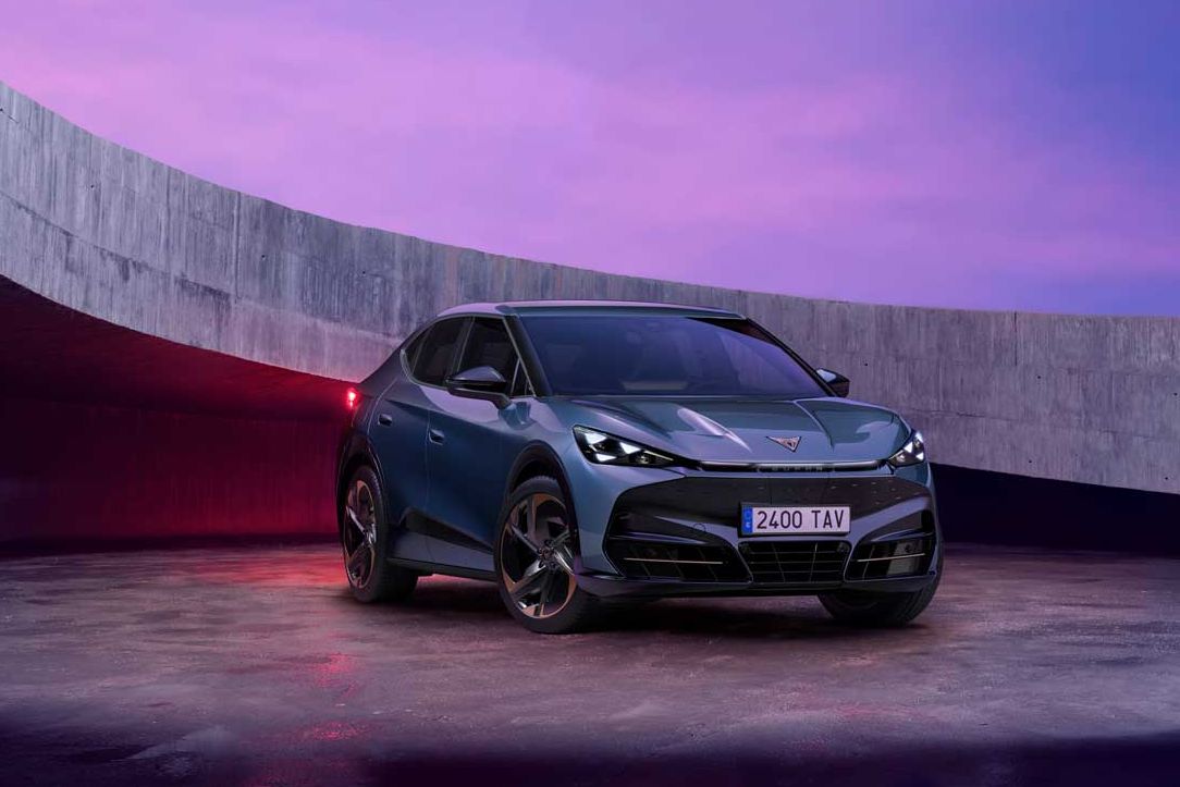 Temen Cupra Tavascan stoji na odprtem prostoru, obdanim z betonskim zidom, ob sončnem zahodu. Avto je športen, temno sive barve, z modernim dizajnom. Nebo je obarvano v vijoličnih in rožnatih odtenkih.