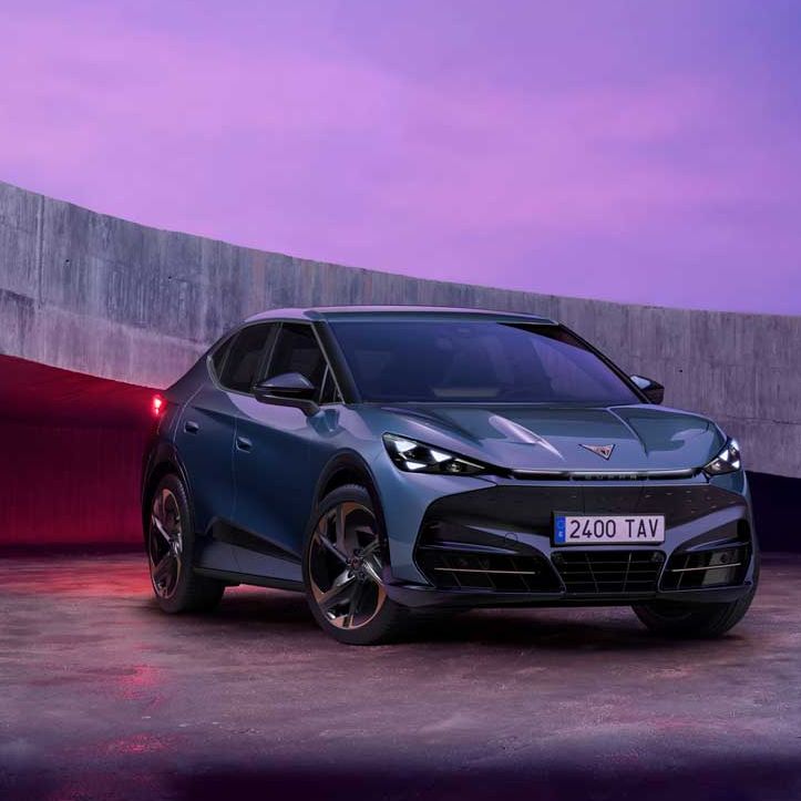 Temen Cupra Tavascan stoji na odprtem prostoru, obdanim z betonskim zidom, ob sončnem zahodu. Avto je športen, temno sive barve, z modernim dizajnom. Nebo je obarvano v vijoličnih in rožnatih odtenkih.