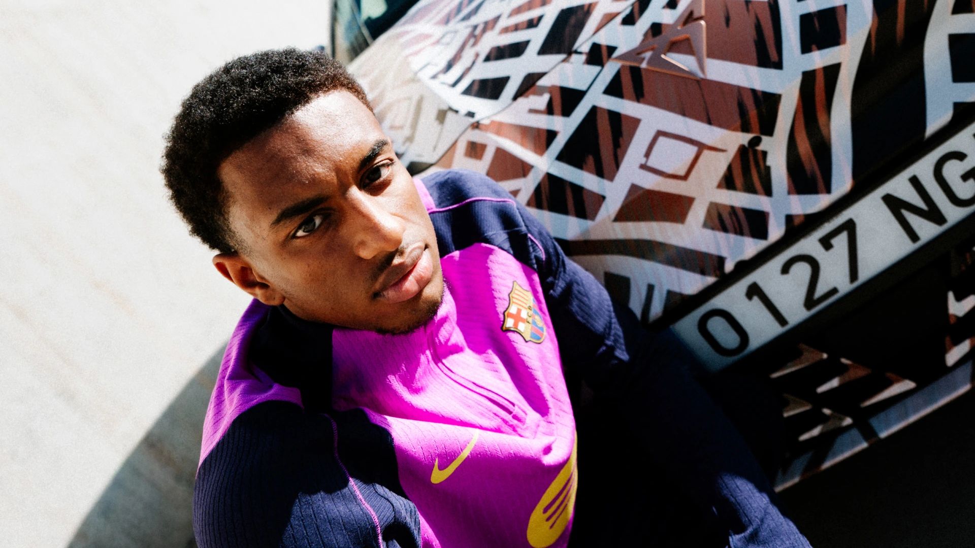 Nogometaš FC Barcelona v roza dresu pozira v počepu ob CUPRA Ravalu. V ozadju napis: BEYOND REBEL, RAVAL. 