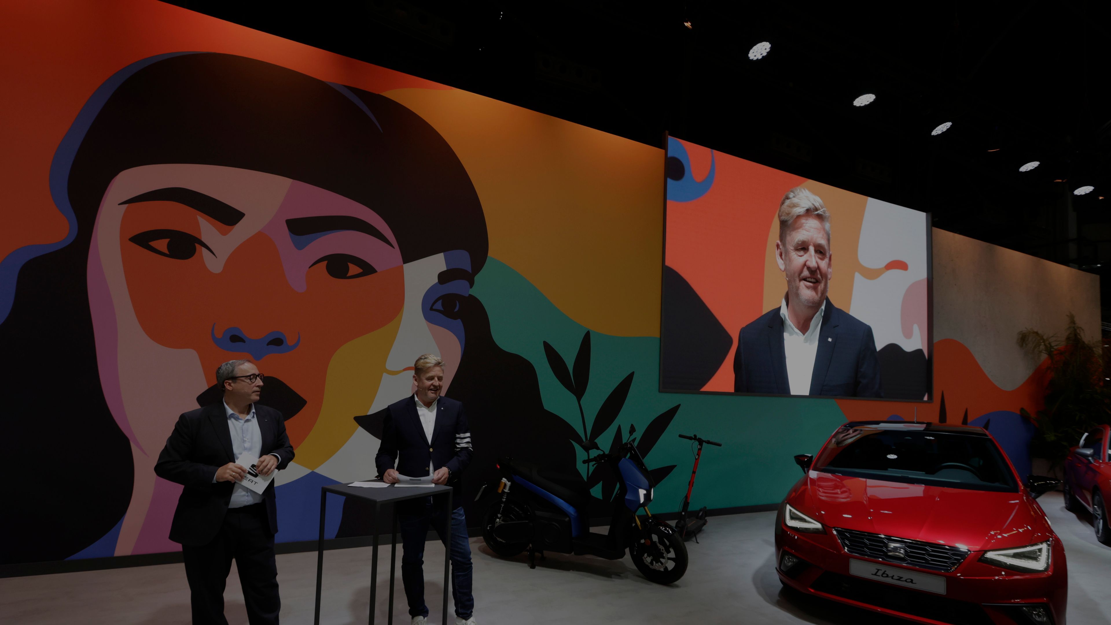 Prostor z barvito steno, na kateri so umetniške slike, vključno z digitalnim zaslonom. Ob steni je rdeče vozilo znamke CUPRA, poleg njega pa motorno kolo. Zraven stoji in drži govor Wayne Griffiths.