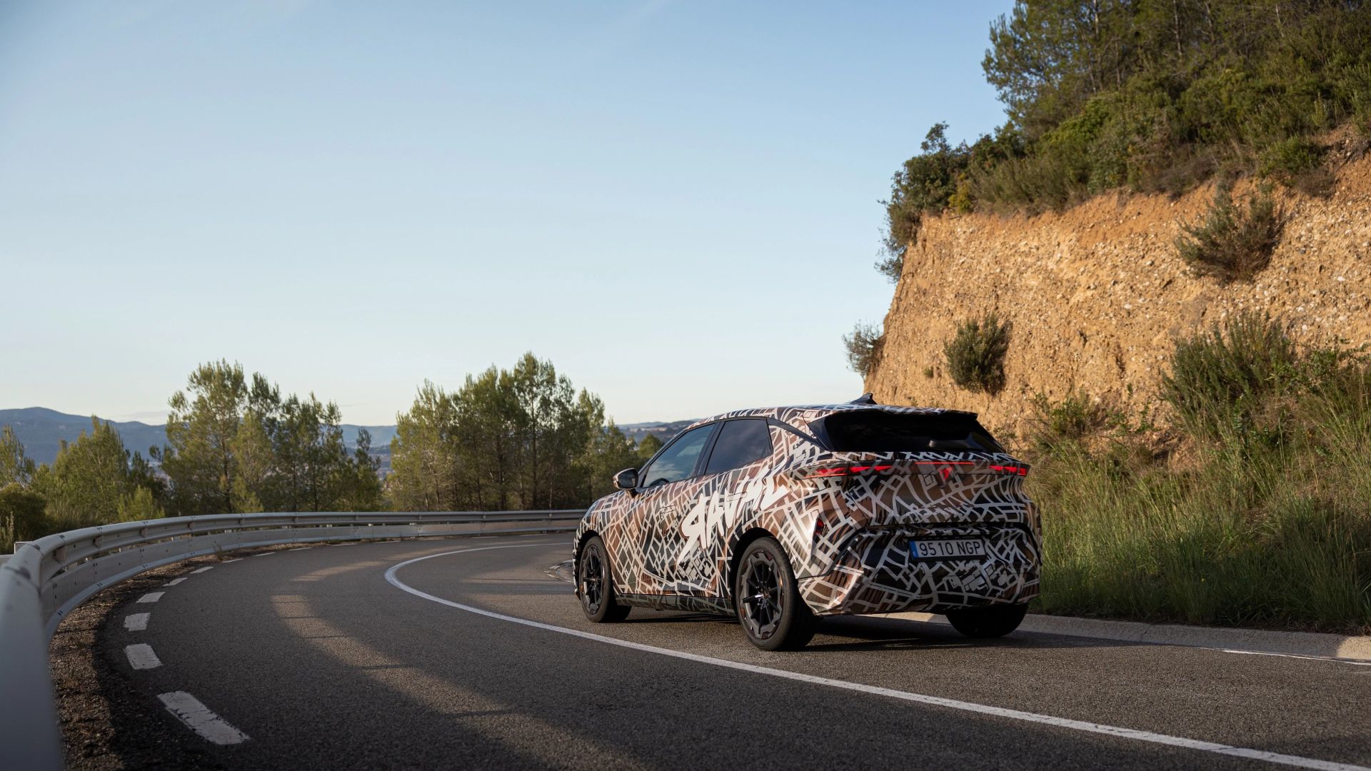 CUPRA Raval Dynamic z maskirnim vzorcem vozi po ovinkasti cesti ob robu hriba. Okoli ceste so drevesa in grmičevje, v daljavi so vidne gore pod jasnim nebom.