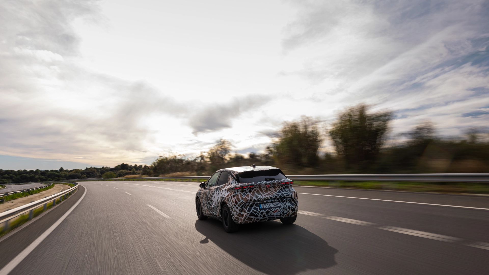 CUPRA Raval Dynamic v maskirni foliji vozi po avtocesti ob sončnem zahodu. Cesta je prazna, ob robu so vidni drevesi. Nebo je delno oblačno.