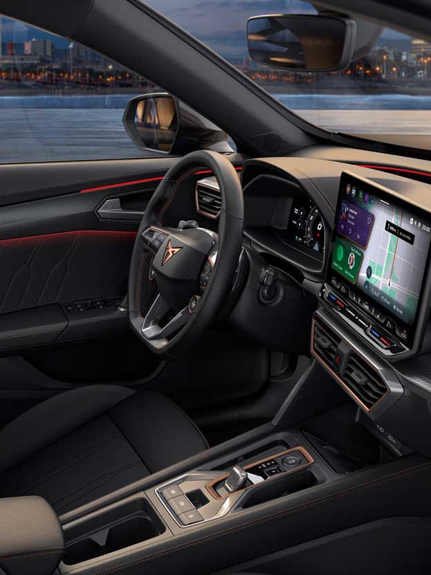 Notranjost sodobnega vozila CUPRA Formentor z velikim zaslonom infotainment sistema, volanom in upravljalnimi elementi. Vozilo ima elegantne črne sedeže, skozi okna pa se vidi osvetljeno mesto.