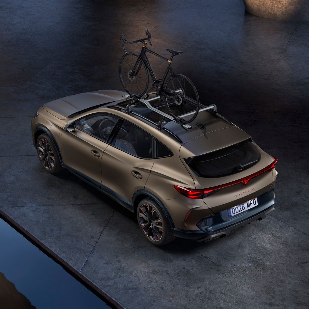 Vozilo CUPRA Formentor PA, parkirano ob vodni površini. Na strehi pa ima nameščeno kolo. Avto je svetlo rjave mat barve z elegantnim dizajnom. Vozilo je temno sivo in ima elegantno, sodobno obliko. Okolica je industrijska, z betonskimi tlemi in stenami.