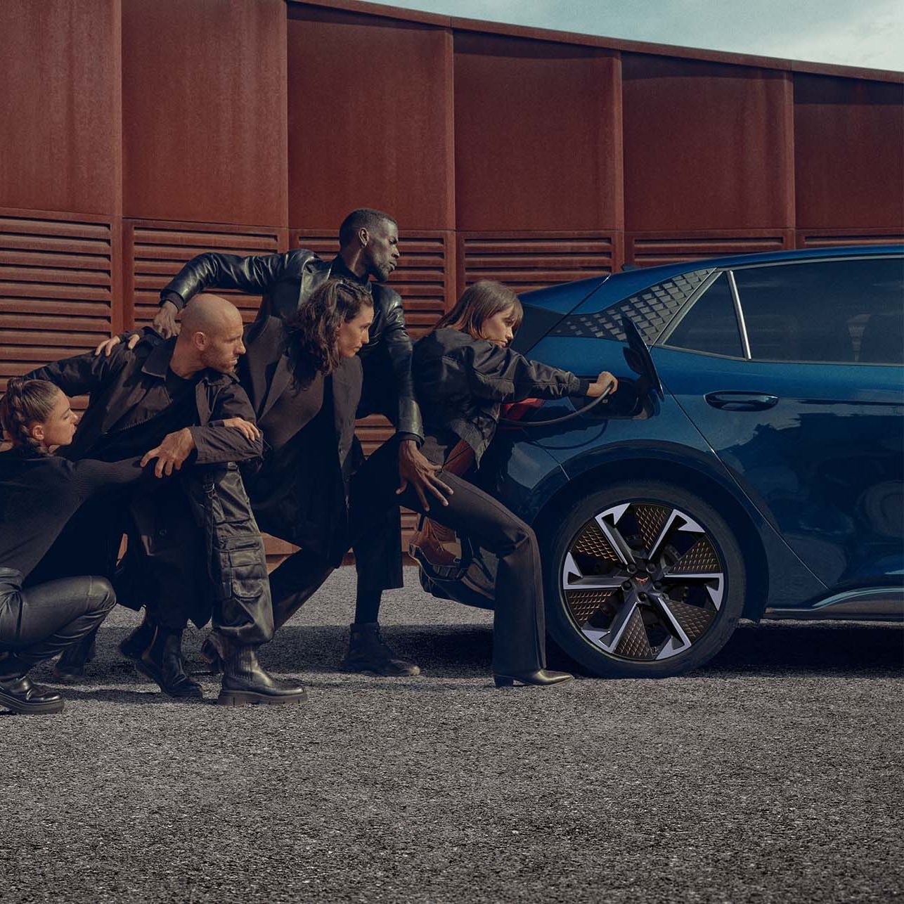 Štirje ljudje v črnih oblačilih stojijo v plesnem položaju zunaj in potiskajo modri električni Cupra Born proti polnilni postaji. Polnilna postaja je sive barve, stena za njimi pa rjave.