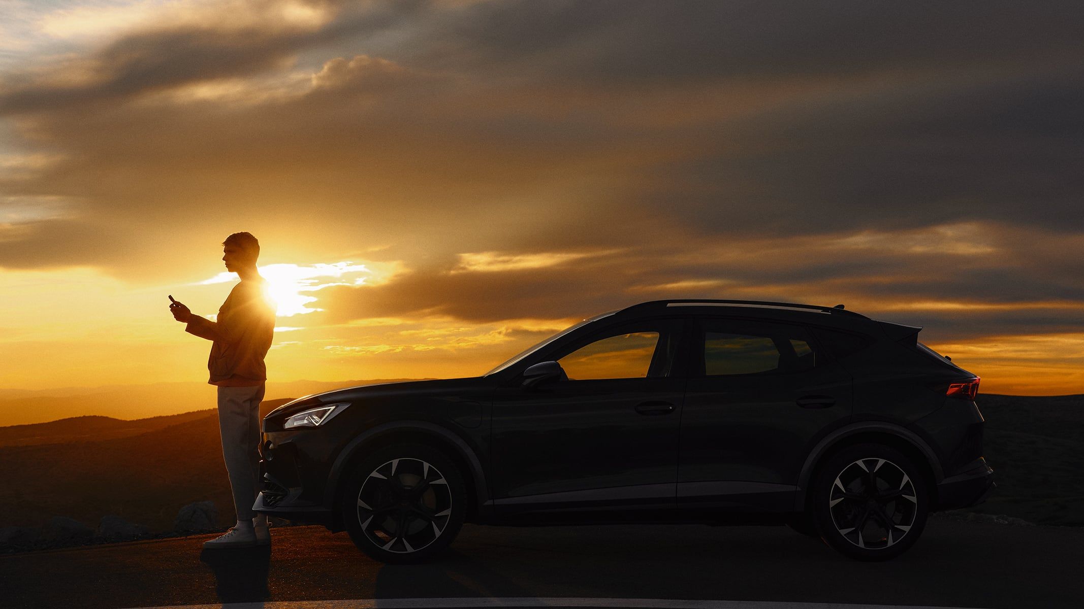 Oseba stoji ob avtomobilu CUPRA Umbrella Formentor, na robu ceste med sončnim zahodom. Nebo je obarvano v oranžne in sive odtenke, ki ustvarjajo dramatično ozadje. 