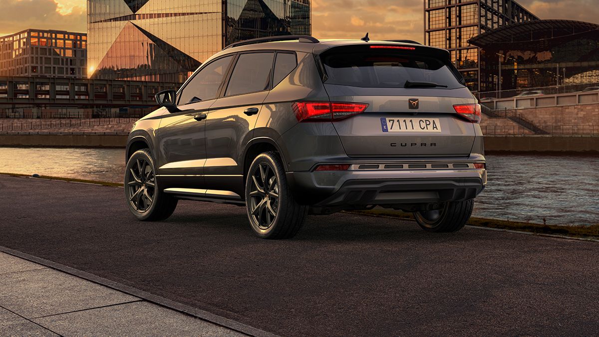 CUPRA Ateca je parkiran ob reki ob sončnem zahodu. V ozadju sta moderna steklena zgradba in industrijsko poslopje. Nebo je obarvano z rumenimi in oranžnimi odtenki.