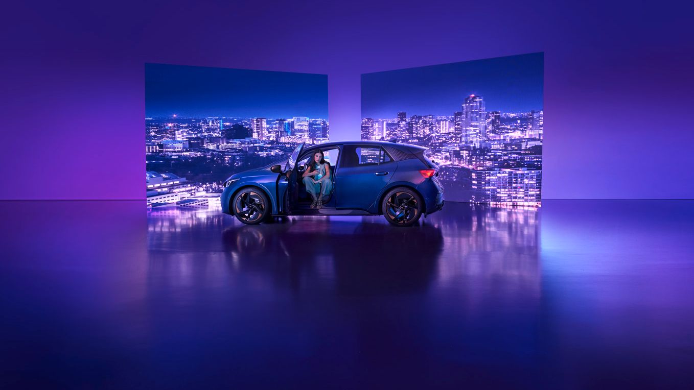 Modra športna CUPRA Born s spredaj odprtimi vrati je parkirana v studiu z vijolično osvetlitvijo. Na sprednjem sedežu sedi ženska, v ozadju pa sta dva velika zaslona z nočno mestno pokrajino.