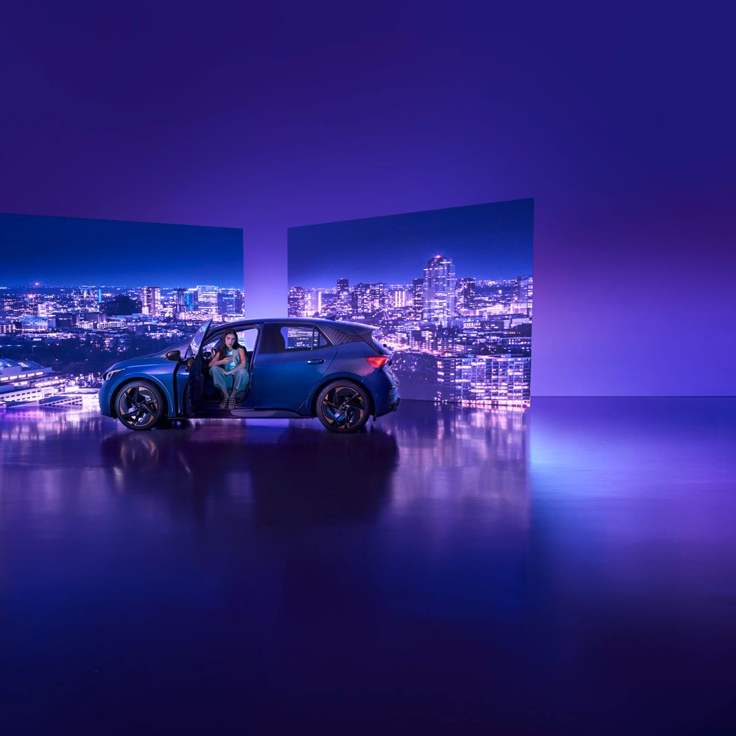 Modra športna CUPRA Born s spredaj odprtimi vrati je parkirana v studiu z vijolično osvetlitvijo. Na sprednjem sedežu sedi ženska, v ozadju pa sta dva velika zaslona z nočno mestno pokrajino.
