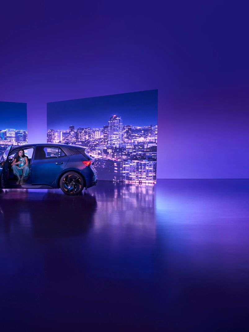 Modra športna CUPRA Born s spredaj odprtimi vrati je parkirana v studiu z vijolično osvetlitvijo. Na sprednjem sedežu sedi ženska, v ozadju pa sta dva velika zaslona z nočno mestno pokrajino.