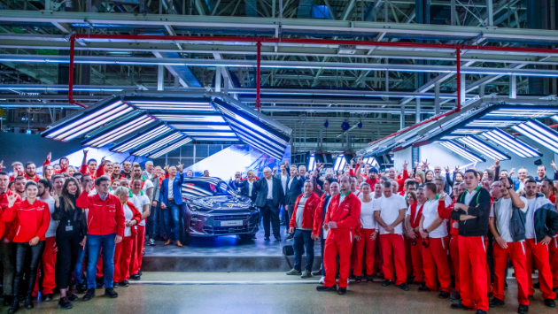 Skupina ljudi, oblečenih v rdeče uniforme, stoji okoli avtomobila znamke CUPRA v proizvodni hali. Skupina vključuje moške in ženske različnih starosti. Območje je dobro osvetljeno z močno industrijsko razsvetljavo.