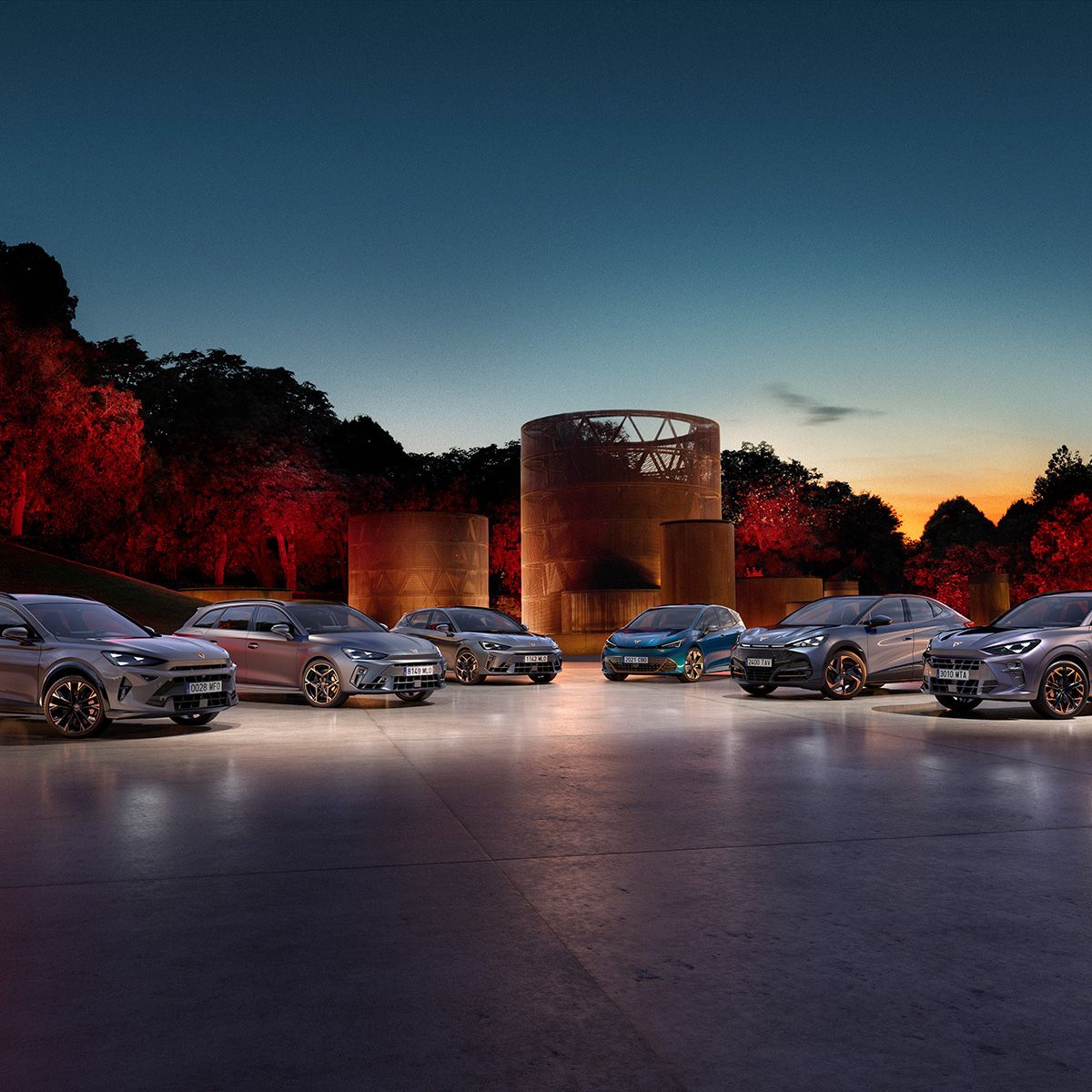 Več vozil CUPRA – CUPRA Born, CUPRA Leon PA 5D, CUPRA Leon PA SP, CUPRA Formentor PA, CUPRA Terramar in CUPRA Tavascan – je razporejenih na odprtem prostoru ob sončnem zahodu. Obkrožajo jih drevesa, osvetljena z rdečo svetlobo, v ozadju pa stoji velik kovinski objekt okrogle oblike.