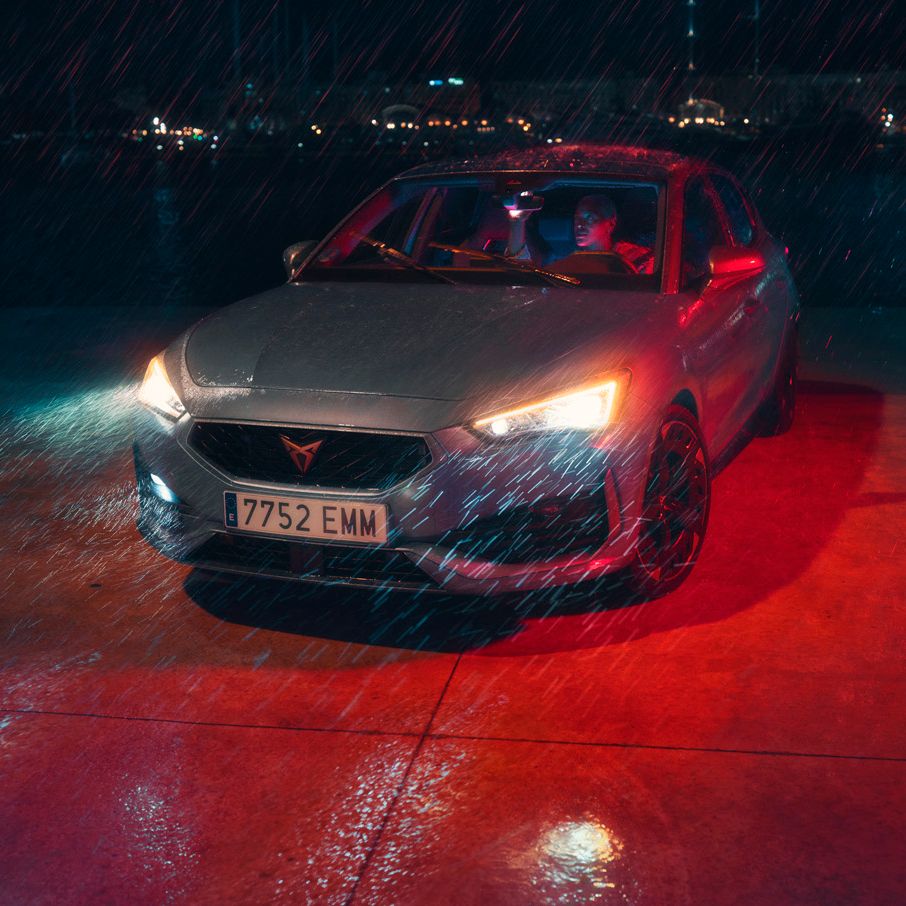 Avtomobil CUPRA Leon PA 5D z vklopljenimi sprednjimi lučmi parkiran na mokrem pločniku v dežju. V ozadju je nočno obzorje z razsvetljenimi stavbami.